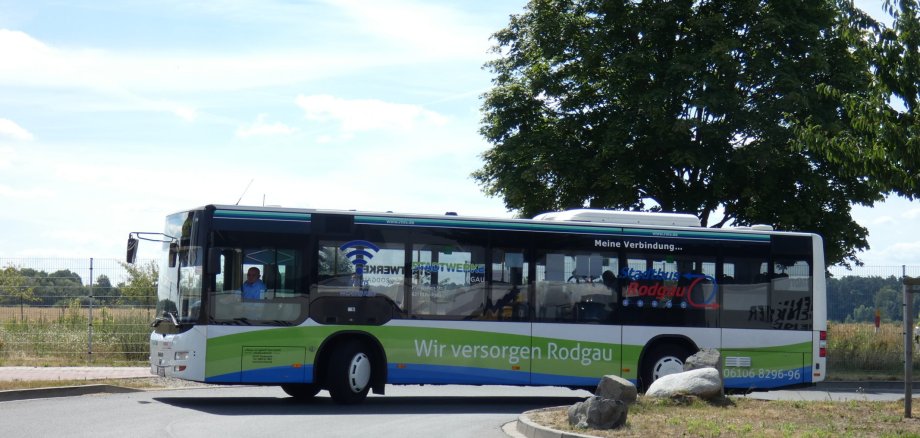 Stadtbus der Linie OF-40 in Rodgau mit Aufschrift 'Wir versorgen Rodgau' fährt auf einer Straße mit Baum und Felsen am Rand