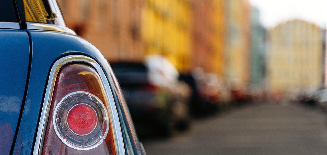 Im Vordergrund ist das Rücklicht eines Autos zu sehen, im Hintergrund eine Straßenschlucht mit Häusern, vor denen viele Autos parken.