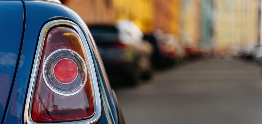 Im Vordergrund ist das Rücklicht eines Autos zu sehen, im Hintergrund eine Straßenschlucht mit Häusern, vor denen viele Autos parken.