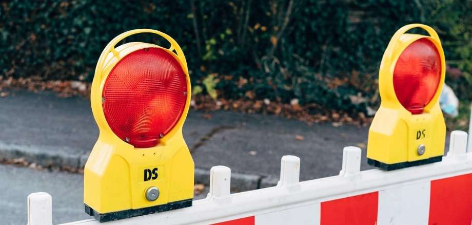 Zwei gelbe Warnleuchten mit rotem Licht auf einer weißen Absperrung mit roten und weißen Streifen. Im Hintergrund sind grüne Pflanzen sichtbar.