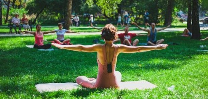 Eine Person in einem rosa Yoga-Outfit sitzt im Schneidersitz auf einer Matte und hat die Arme seitlich ausgestreckt. Im Hintergrund sind mehrere Personen ebenfalls beim Yoga auf Matten.