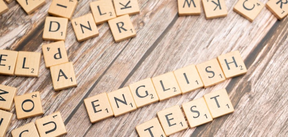 Holzspielsteine mit Buchstaben, die das Wort 'ENGLISH TEST' bilden, umgeben von weiteren Buchstaben auf einem Holztisch.