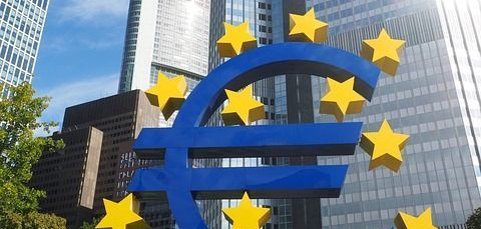 Eine große, blaue Euro-Skulptur mit gelben Sternen steht im Vordergrund. Im Hintergrund sind moderne Hochhäuser und ein blauer Himmel zu sehen.