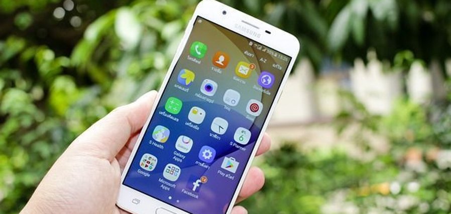 Ein weißes Samsung-Smartphone in einer Hand, mit einem bunten App-Layout auf dem Bildschirm, im Hintergrund sind grüne Pflanzen sichtbar.