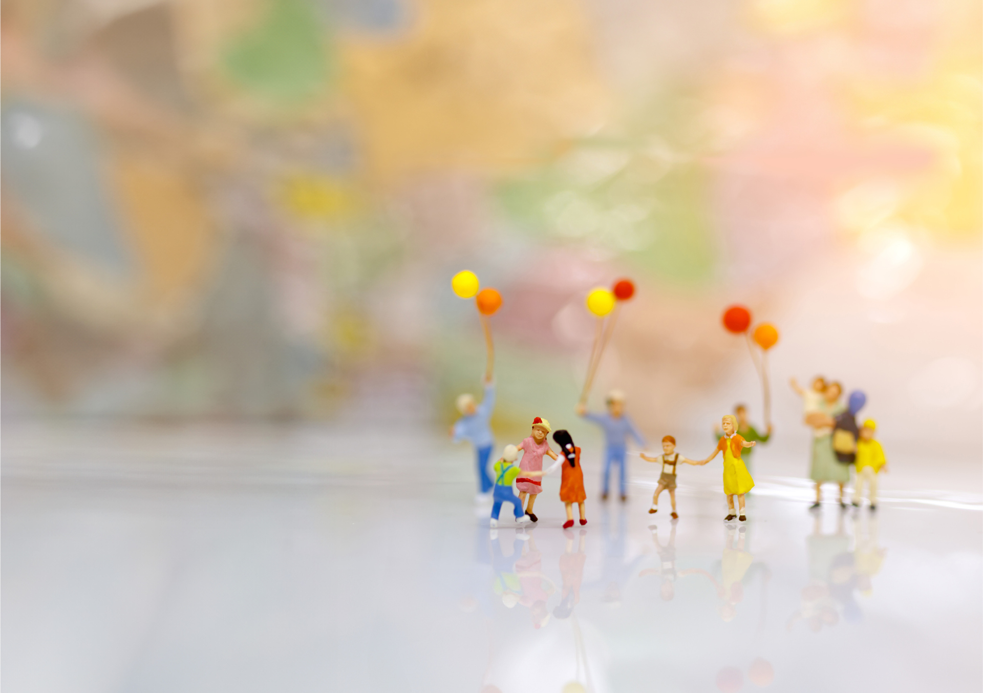 Symbolbild der Miteinander-Zeit Gruppe winziger Figuren mit bunten Luftballons auf hellem Untergrund vor unscharfem, farbigem Hintergrund