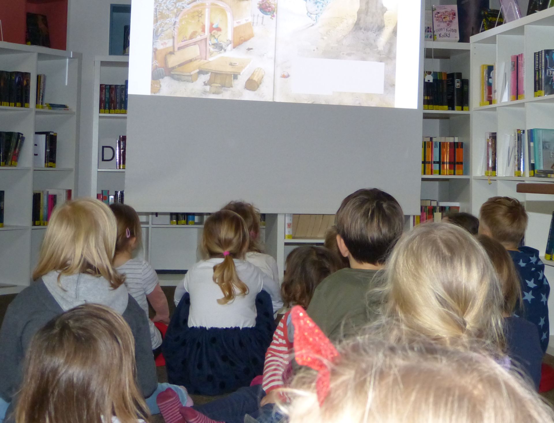 Bilderbuchkino Kinder sitzen auf dem Boden in einer Bibliothek und schauen ein Bilderbuchkino mit einer gezeichneten Szene an.