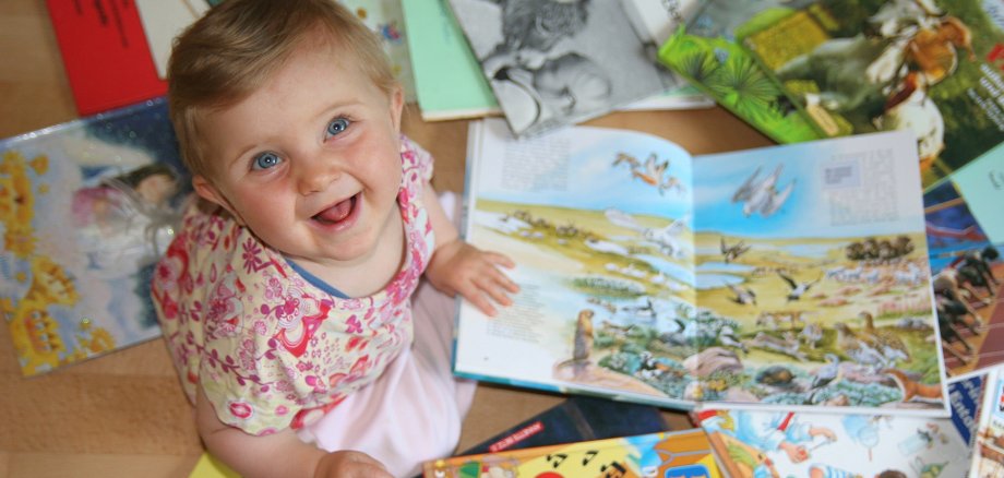 Kleines Kind sitzt auf dem Boden und schaut sich mehrere aufgeschlagene Kinderbücher mit bunten Illustrationen an