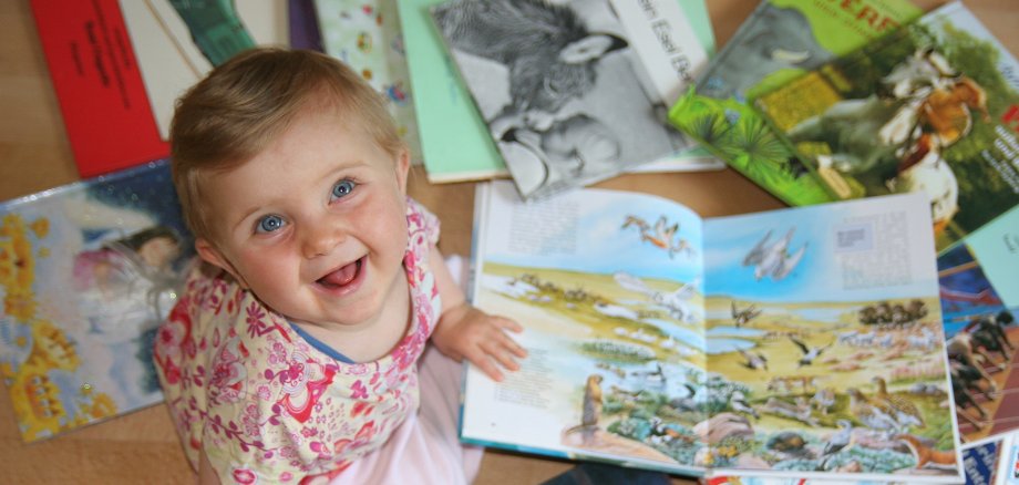 Kleines Kind sitzt auf dem Boden und schaut sich mehrere aufgeschlagene Kinderbücher mit bunten Illustrationen an