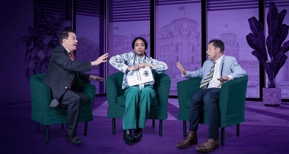 Drei Personen sitzen in grünen Sesseln in einem Studio, zwei Männer in Anzügen diskutieren mit einer Frau in medizinischer Kleidung, die ein Notizbuch hält