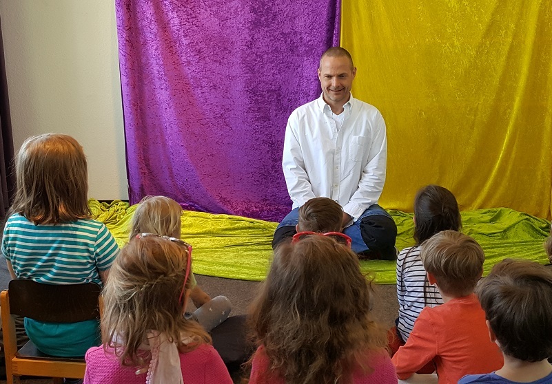Sitzkreis Kinder Ein Erwachsener sitzt auf dem Boden vor einem lila und gelben Vorhang und erzählt einer Gruppe von Kindern, die auf Stühlen und dem Boden sitzen und ihm zuhören.