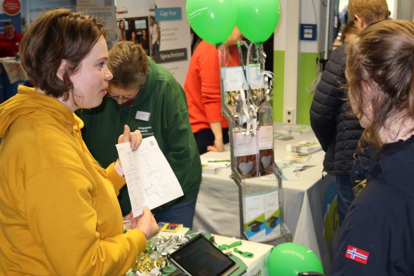 Besucherinnen und Besucher an einem Messestand mit Informationsmaterial und grünen Luftballons auf einer Bildungsmesse