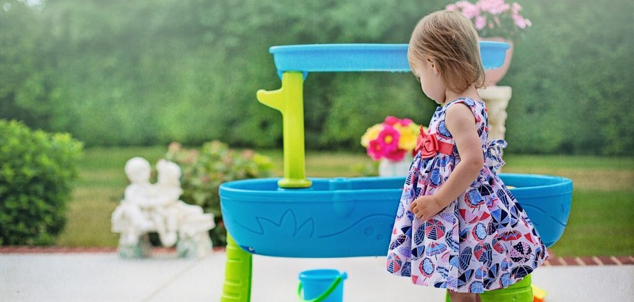 Kleines Mädchen in gemustertem Kleid und Sandalen steht vor einem blauen und grünen Spieltisch mit Eimer im Garten