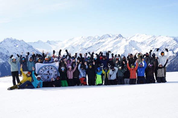 Große Gruppe von Menschen in Skibekleidung posiert mit erhobenen Armen vor verschneiten Bergen beim Snowcamp 2024