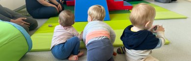 Drei Kleinkinder Drei Kleinkinder sitzen und krabbeln auf einer hellgrünen Matte. Im Hintergrund sind weitere Kinder und Erwachsene zu sehen, die um eine blaue und rote Spielstruktur sitzen. Die Kinder tragen unterschiedliche, farbige Kleidung, darunter gestreifte Socken und Pullover. Spielzeug liegt auf der Matte verteilt.