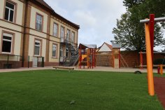 Spielplatz mit Klettergerüst und Rutsche neben einem zweistöckigen Schulgebäude mit Außenfluchttreppe