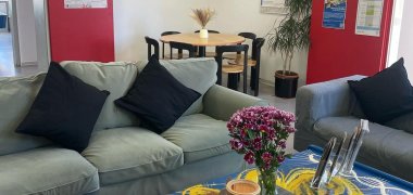 Im Vordergrund sind zwei Sofas davor ein Tisch, auf dem Blumen stehen. Im Hintergrund befindet sich eine Tisch mit Stühlenrundrum.
