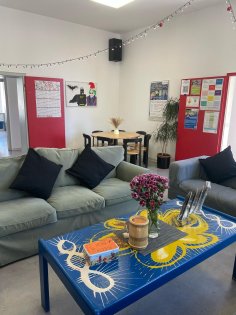 Jugendtreff Jügesheim Im Vordergrund sind zwei Sofas davor ein Tisch, auf dem Blumen stehen. Im Hintergrund befindet sich eine Tisch mit Stühlenrundrum.