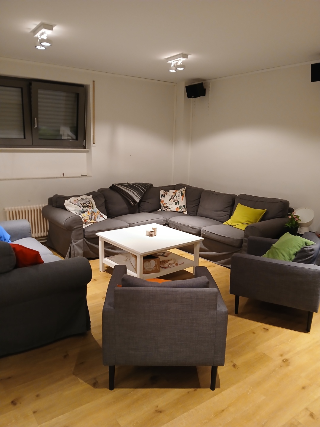 Es steht ein graues Ecksofa, sowie drei Sessel in diesem Raum. Auf diesen liegen viele bunte Kissen und es steht ein weißer Tisch dabei.