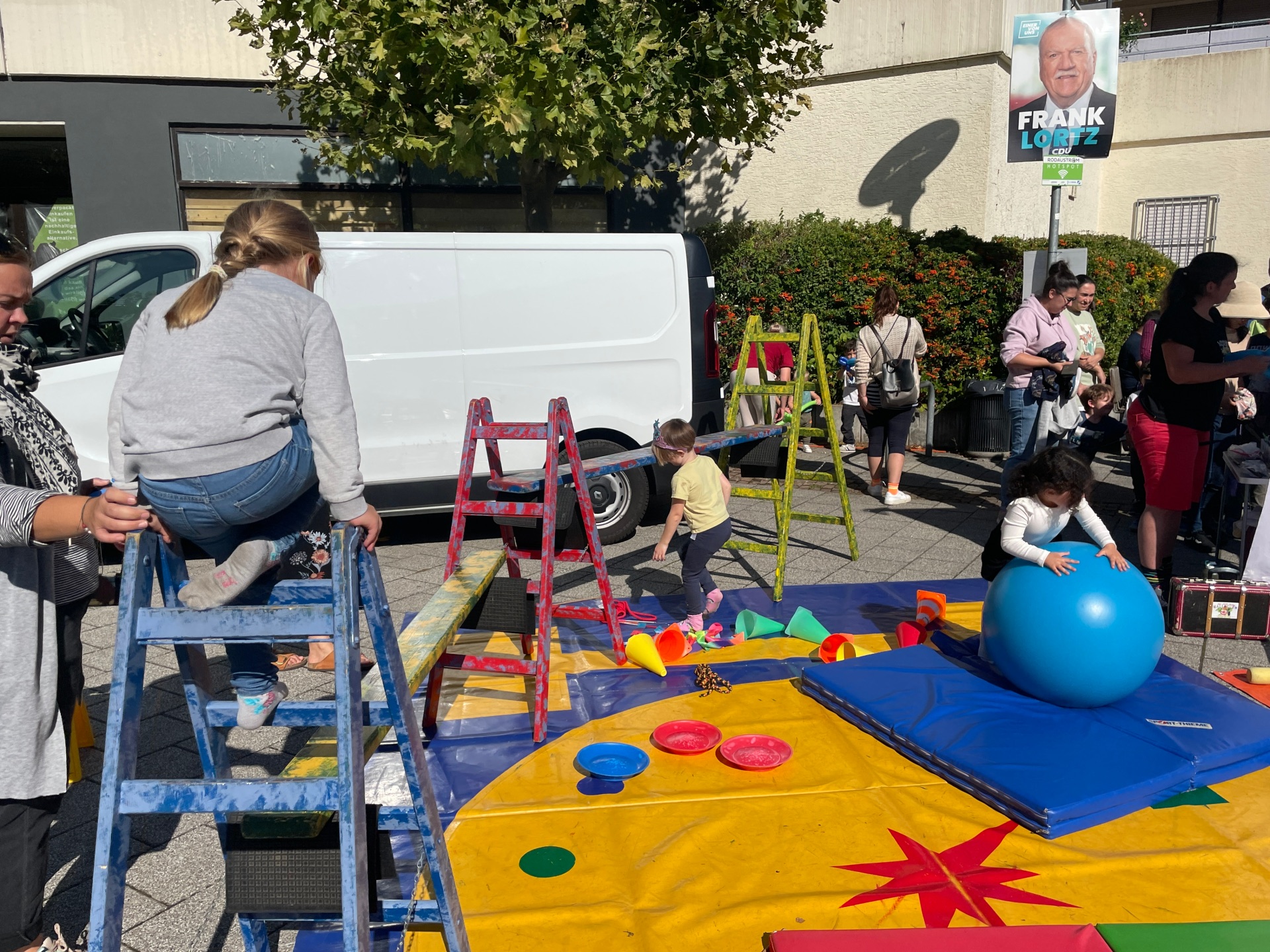 Kinder spielen auf einem bunten Spielplatz mit Leitern, Matten und einem großen blauen Gymnastikball bei einem Fest im Freien