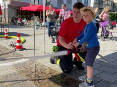 Kind mit Strohhut hält Feuerwehrschlauch, unterstützt von Feuerwehrmann in rotem T-Shirt, bei Kinderfest im Freien