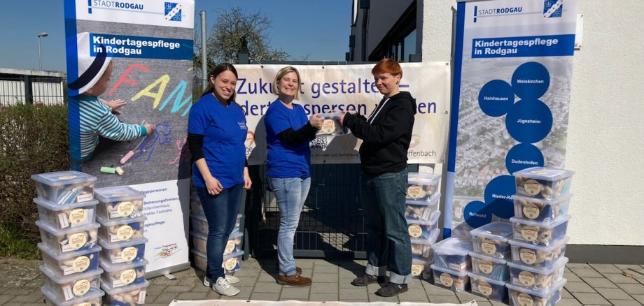 Drei Personen vor Bannern der Stadt Rodgau Drei Personen vor Bannern der Stadt Rodgau informieren über Kindertagespflege, mit Stapeln von Kisten mit Spielzeug neben ihnen