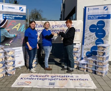 Drei Personen vor Bannern der Stadt Rodgau Drei Personen vor Bannern der Stadt Rodgau informieren über Kindertagespflege, mit Stapeln von Kisten mit Spielzeug neben ihnen
