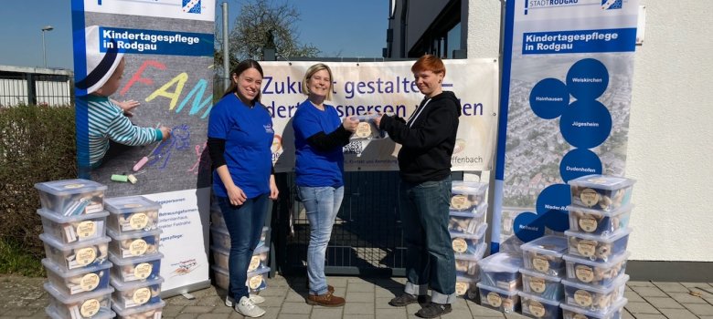 Drei Personen vor Bannern der Stadt Rodgau Drei Personen vor Bannern der Stadt Rodgau informieren über Kindertagespflege, mit Stapeln von Brotdosen neben ihnen