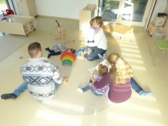 Drei Erwachsene und drei Kinder sitzen auf dem Boden eines hellen Gruppenraums einer Kita, spielen und lesen gemeinsam