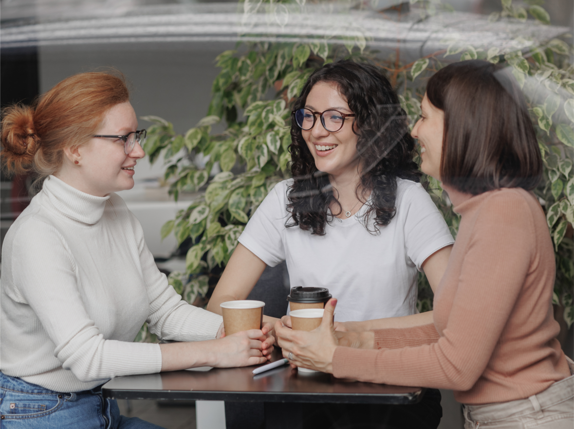 Drei Frauen sitzen an einem Tisch, halten Pappbecher und unterhalten sich, im Hintergrund eine grüne Pflanze