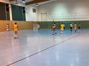Jugendliche spielen Hallenfußball in einer Sporthalle, einige sitzen auf der Bank und schauen zu