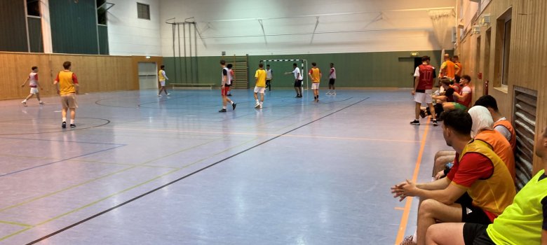 Jugendliche spielen Hallenfußball in einer Sporthalle, einige sitzen auf der Bank und schauen zu
