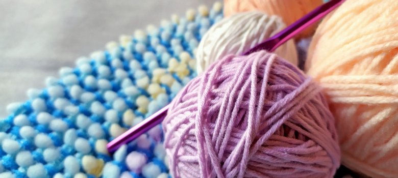 Drei Wollknäuel in verschiedenen Farben (lila, weiß, orange) liegen auf einem blauen, strukturierten Untergrund mit einer Häkelnadel in lila.