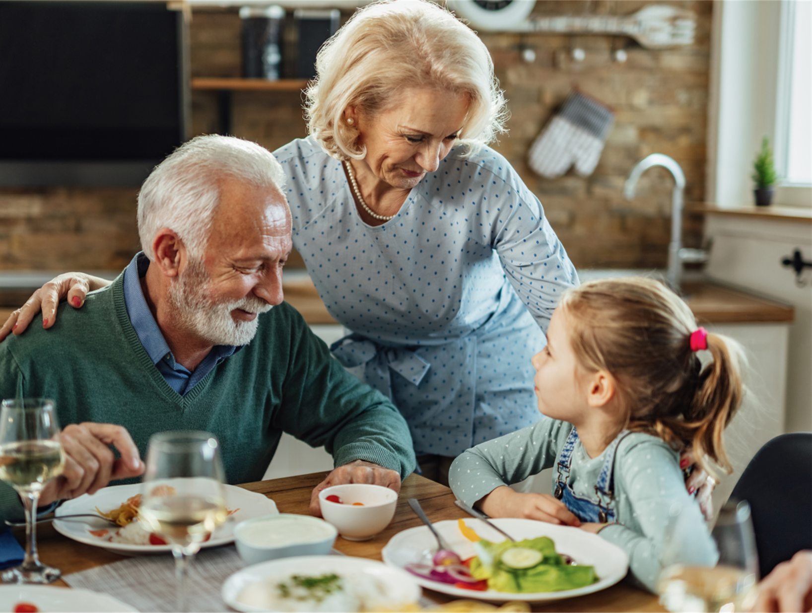 Älteres Paar sitzt am Esstisch, spricht mit einem kleinen Mädchen mit Pferdeschwanz, gedeckter Tisch mit Salat und Getränken