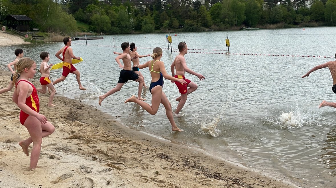Mehrere Kinder und Jugendliche in Badebekleidung laufen ins Wasser des Badesees bei der Eröffnung