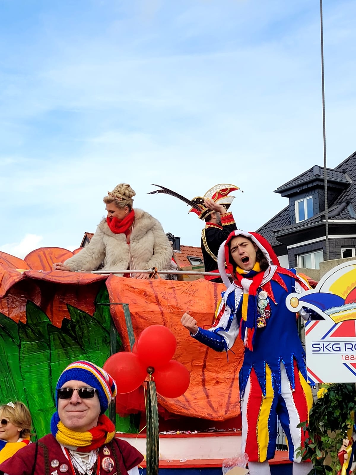 Fastnacht-Prinzenpaar in bunten Kostümen mit Narrenkappe und Feder auf einem Festwagen, umgeben von roten Luftballons und Zuschauern.
