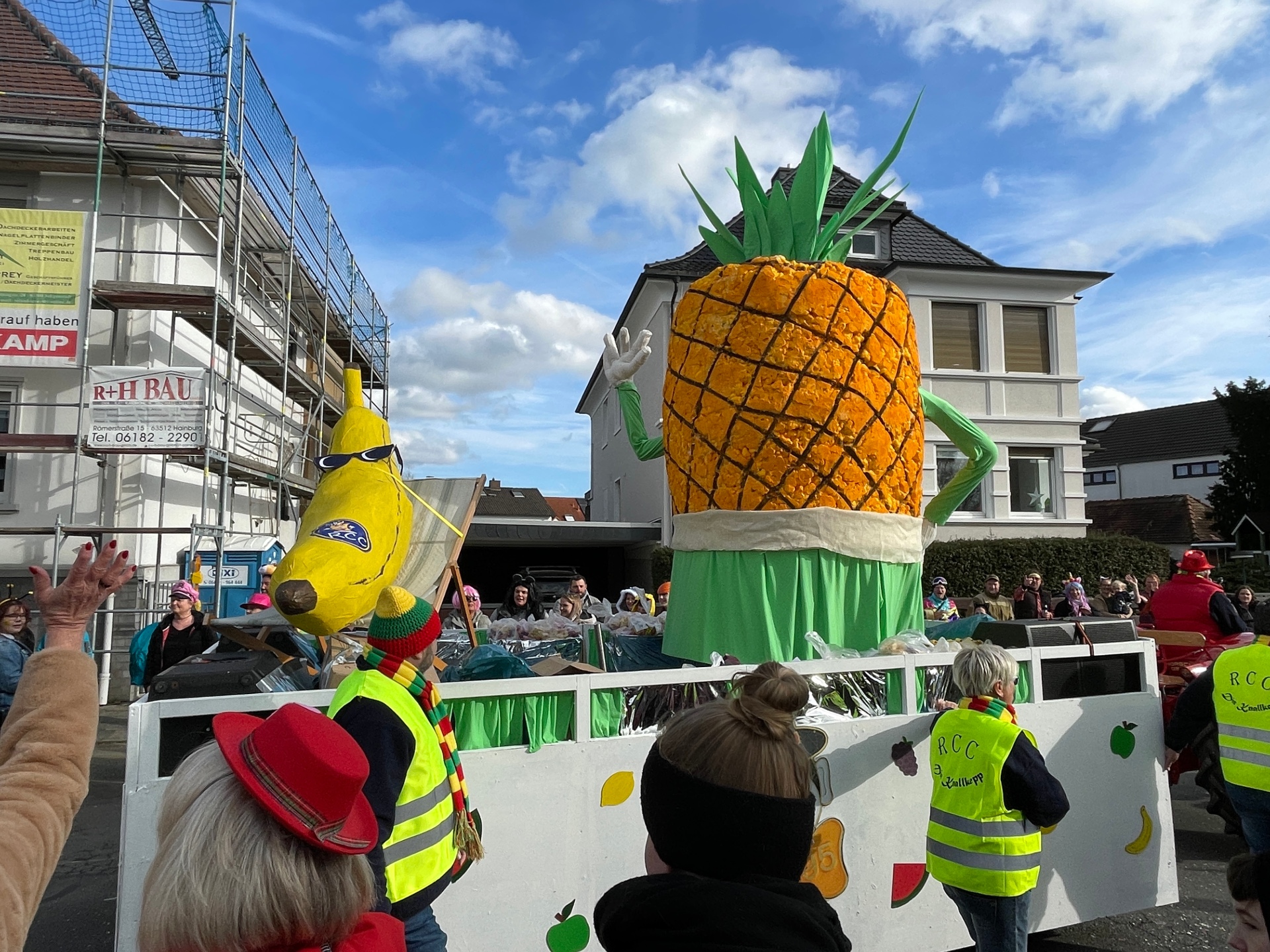 Große Ananas-Figur mit grünen Armen und Blättern auf einem Festwagen vom RCC beim Fastnachtsumzug 2024, umgeben von Sicherheitspersonen in gelben Warnwesten mit 'RCC' Schriftzug.