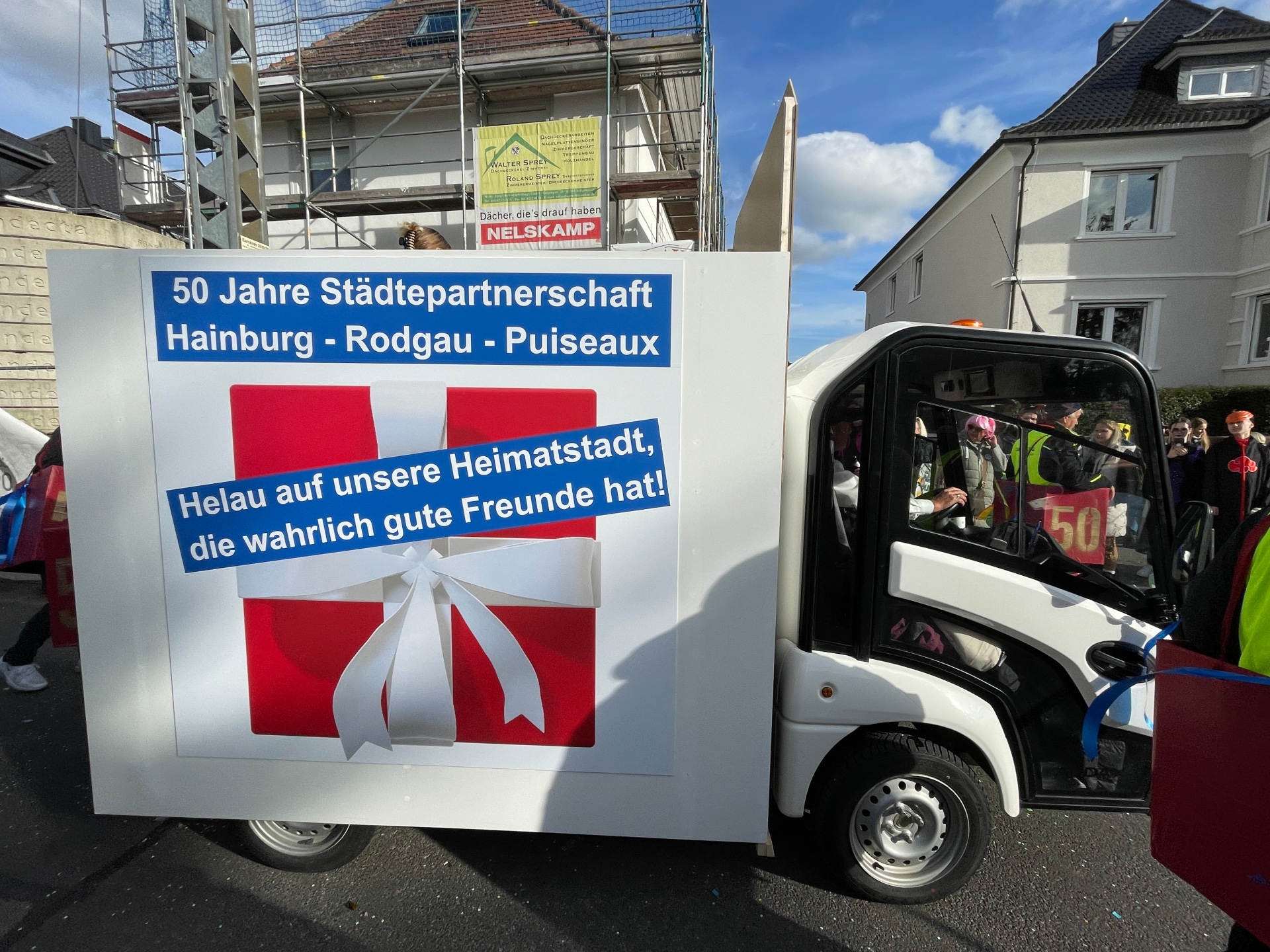 Plakat an einem kleinen weißen Fahrzeug mit Text zu 50 Jahren Städtepartnerschaft Hainburg, Rodgau, Puiseaux und einem Gruß 'Helau auf unsere Heimatstadt, die wahrlich gute Freunde hat!'