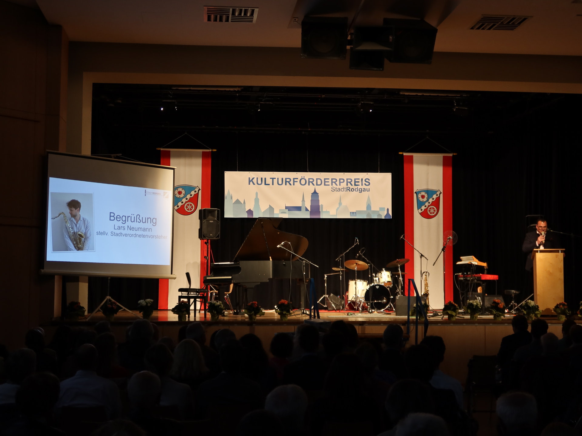 Bühne mit Klavier, Schlagzeug und Mikrofonen bei der Verleihung des Kulturförderpreises Stadt Rodgau, links Leinwand mit Begrüßung von Lars Neumann