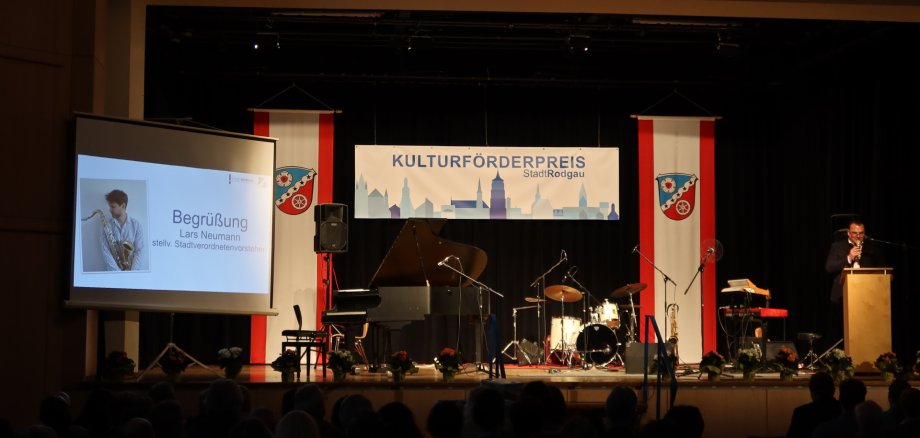 Bühne mit Klavier, Schlagzeug und Mikrofonen bei der Verleihung des Kulturförderpreises Stadt Rodgau, links Leinwand mit Begrüßung von Lars Neumann