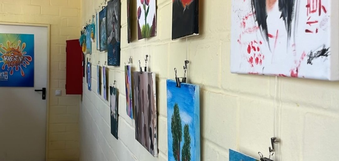 Kunstwerke von der JugendArt hängen an einer weißen Wand in einem Flur, darunter ein Porträt einer Frau mit schwarzem Haar und rotem Hintergrund