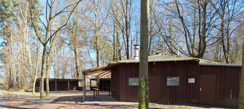 Waldfreizeitanlage Hainhausen Rechts ist das Bewirtschaftungsgebäude der Waldfreizeitanlage Hainhausen zu sehen, im Hintergrund sind der überdachte Sitzbereich und Vorplatz sowie Bäume des Waldes.