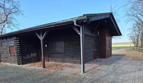 Links ist das Holzgebäude an der  Waldfreizeitanlage Weiskirchen mit gepflastertem Vorplatz, rechts blickt man auf das Feld und etwas Wald.