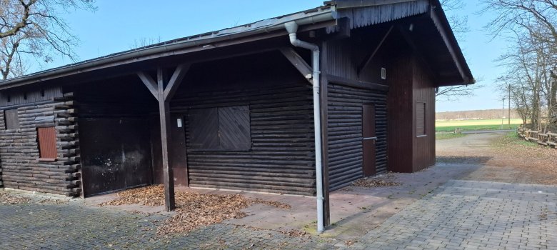 Waldfreizeitanlage Weiskirchen Links ist das Holzgebäude an der Waldfreizeitanlage Weiskirchen mit gepflastertem Vorplatz, rechts blickt man auf das Feld und etwas Wald.