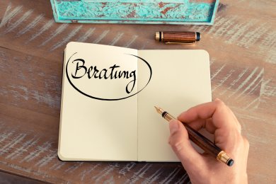 Hand schreibt mit Füllfederhalter das Wort 'Beratung' in ein aufgeschlagenes Notizbuch auf einem Holztisch.