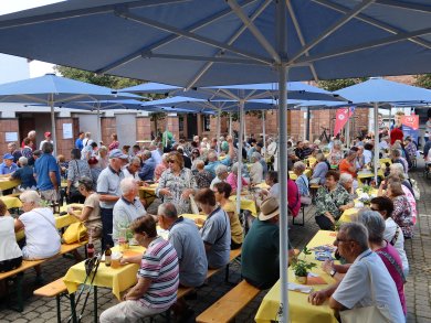 Viele ältere Menschen bei einer Veranstaltung Viele ältere Menschen sitzen unter blauen Sonnenschirmen an langen Holztischen mit gelben Tischdecken bei einer Veranstaltung im Freien