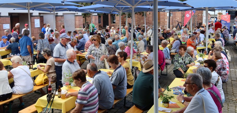Viele ältere Menschen sitzen unter blauen Sonnenschirmen an langen Holztischen mit gelben Tischdecken bei einer Veranstaltung im Freien