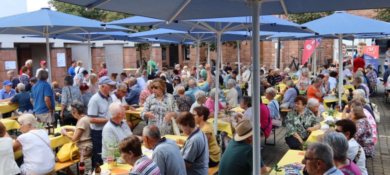 Viele ältere Menschen bei einer Veranstaltung Viele ältere Menschen sitzen unter blauen Sonnenschirmen an langen Holztischen mit gelben Tischdecken bei einer Veranstaltung im Freien