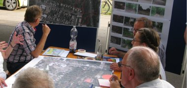 Beteiligung im Rahmen der Bauleitplanung Um einen Tisch, in dessen Mitte ein Plan ist, sitzen Interessierte und Verantwortliche im Dialog.