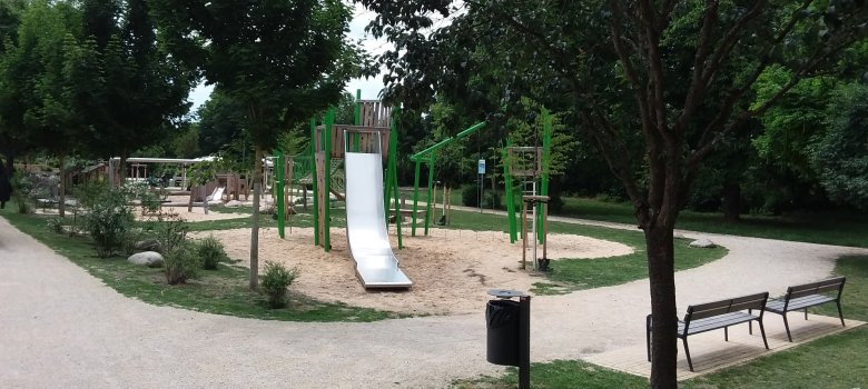 Rodaupark Zwei helle Wege rahmen den Spielbereich mit Rutsche und Klettergerüsten ein. Am Rand sind Bäume und eine Wiese.