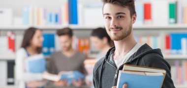 Junger Public Management Student in Bibliothek hält Bücher im Arm und lächelt